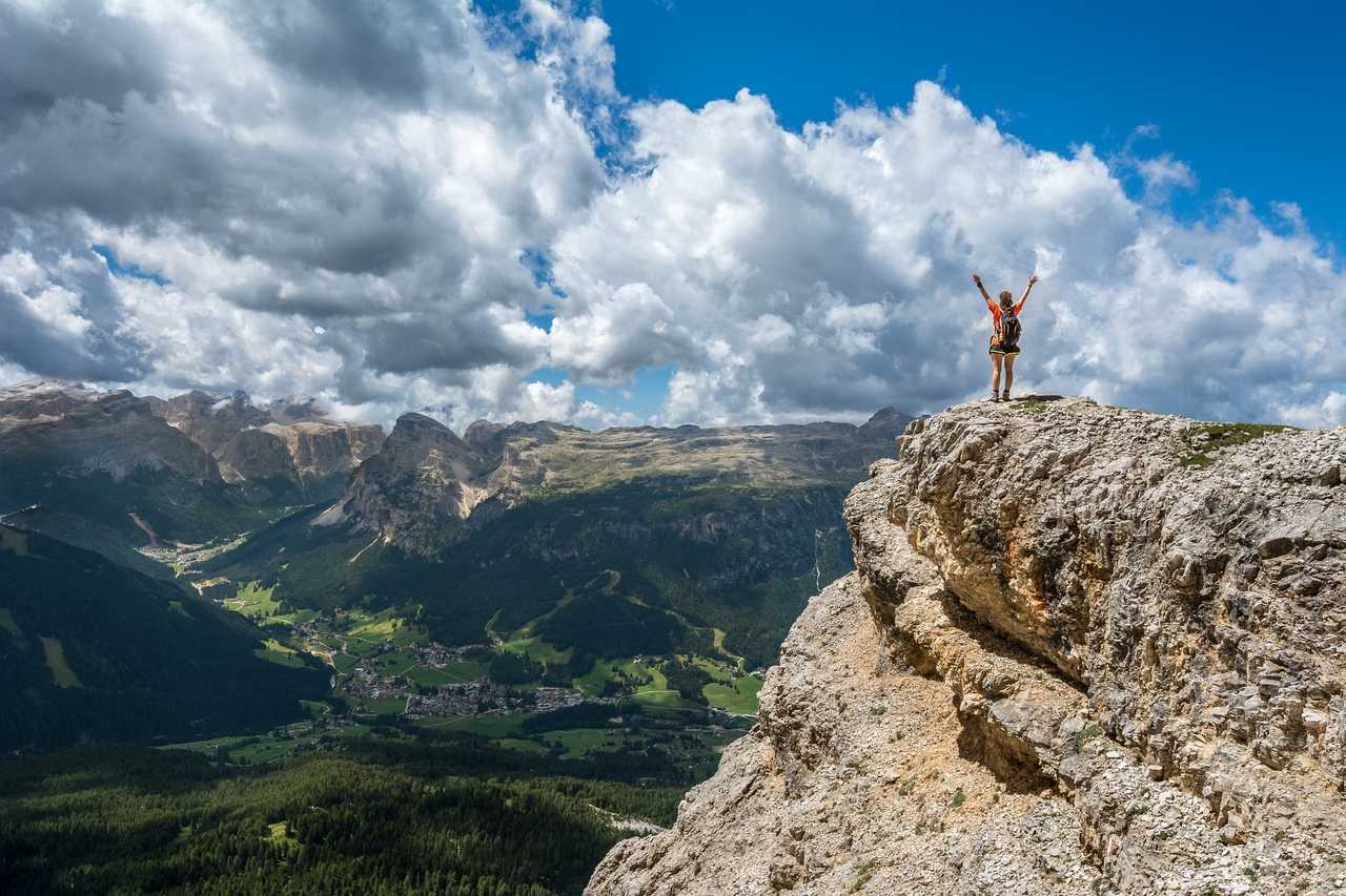 A person stands triumphantly on the edge of a rocky cliff with arms raised, overlooking a vast mountainous landscape with green valleys and a village below